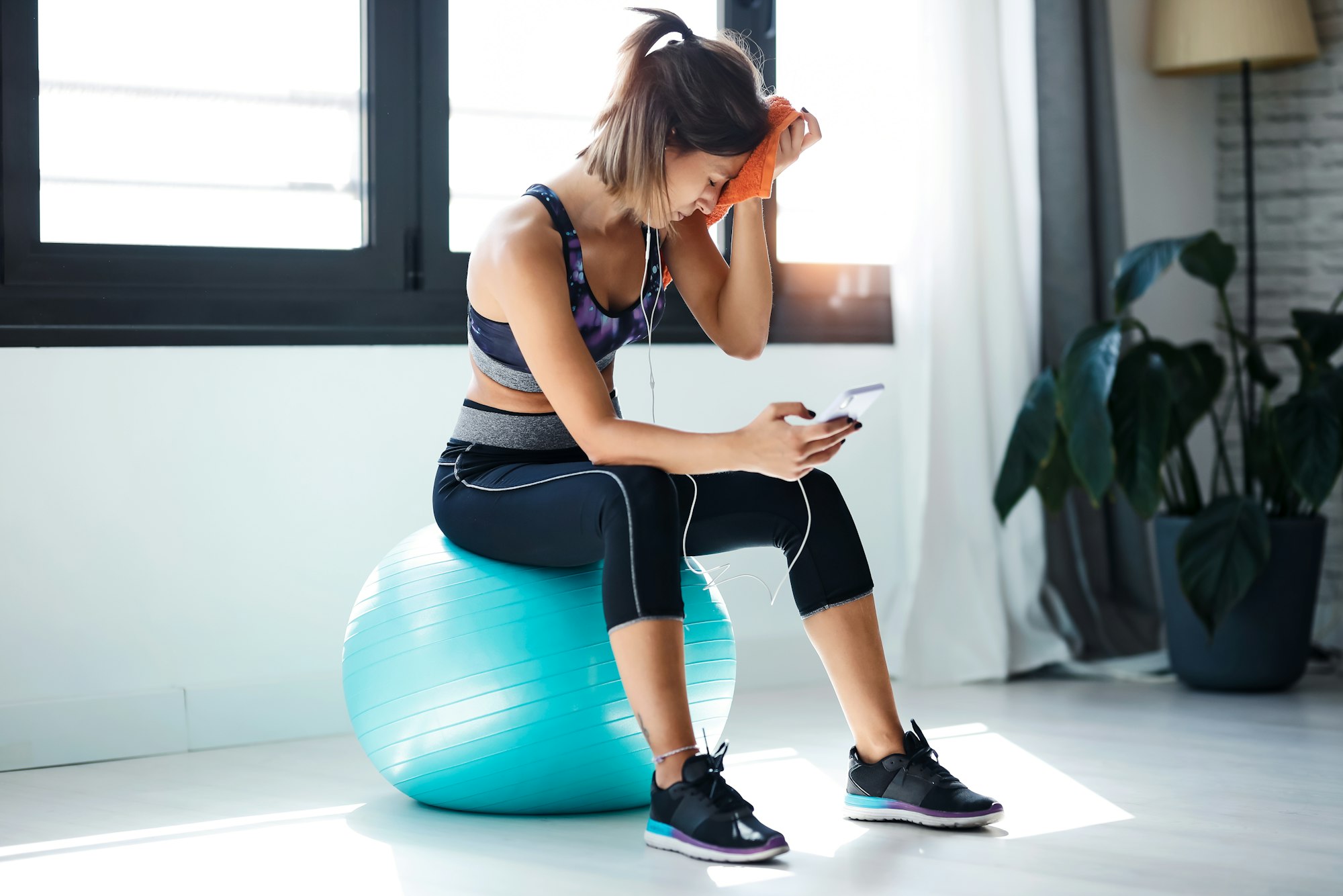 Sporty woman using mobile phone after session of exercises while sitting on fitness ball at home.